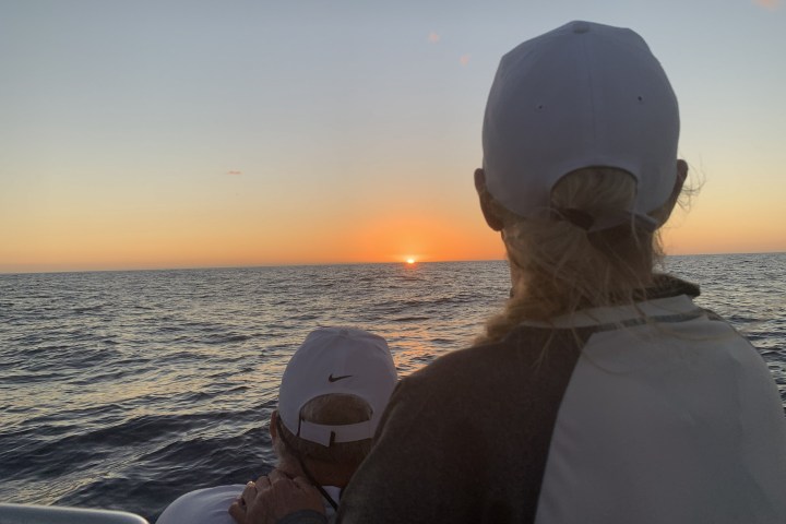 Two people in caps watch a sunset over the ocean from a boat.